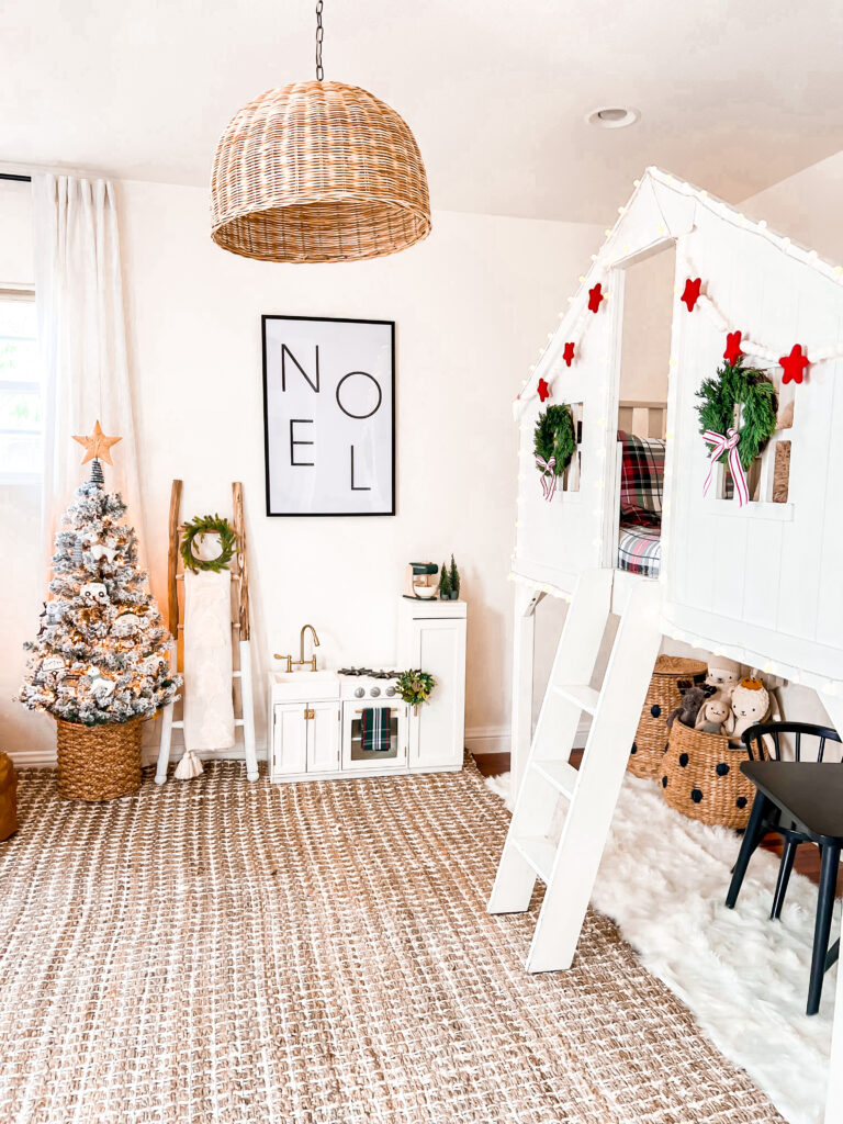 This is a child bedroom decorated for Christmas. There is a brown and white striped large area rug, a white Pottery Barn Kid's loft bed, and a small white child play kitchen with a mini green wreath and green toy mixer. There is a large black framed print that says "Noel." Next to the play kitchen is a wood ladder with a white throw and green mini Christmas wreath. There is also a small flocked Christmas tree with a rattan star on top.