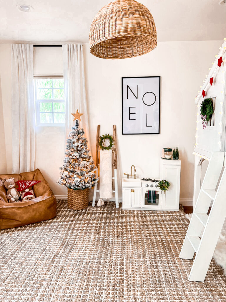 This is a child bedroom decorated for Christmas. There is a brown and white striped large area rug, a white Pottery Barn Kid's loft bed, and a small white child play kitchen with a mini green wreath and green toy mixer. There is a large black framed print that says "Noel." Next to the play kitchen is a wood ladder with a white throw and green mini Christmas wreath. There is also a small flocked Christmas tree with a rattan star on top.