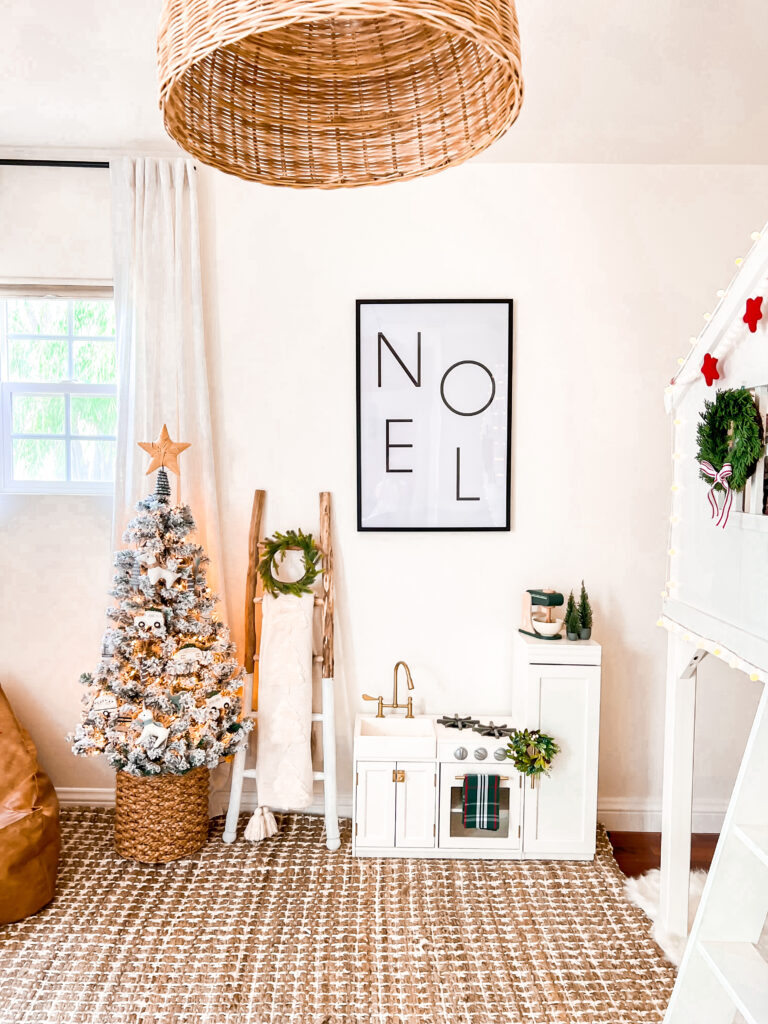 Small white child play kitchen with a mini green wreath and green toy mixer. There is a large black framed print that says "Noel." Next to the play kitchen is a wood ladder with a white throw and green mini christmas wreath. There is also a small flocked Christmas tree with a rattan star on top.