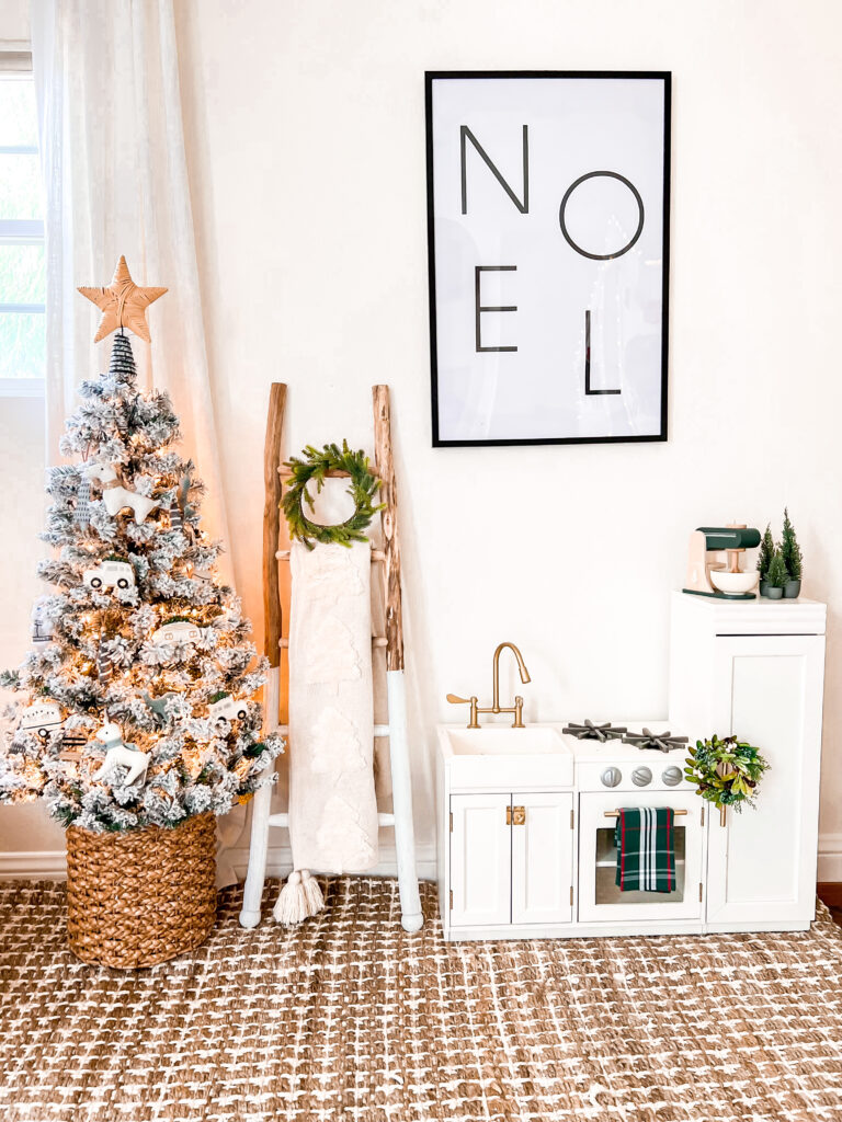 In a bedroom, there is a small white Pottery Barn Kid's play kitchen with a mini green wreath and green toy mixer. There is a large black framed print that says "Noel." Next to the play kitchen is a wood ladder with a white throw and green mini christmas wreath. There is also a small flocked Christmas tree with a rattan star on top.