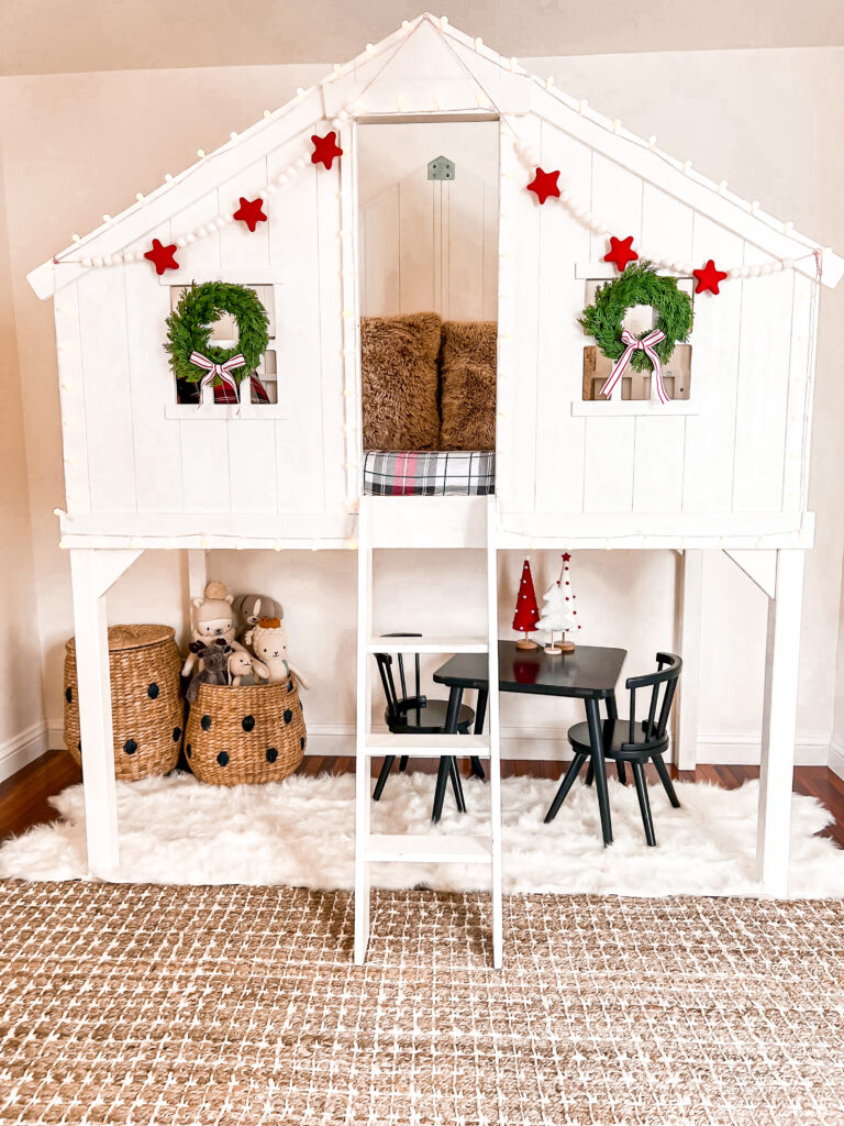 This is a child bedroom decorated for Christmas. There is a brown and white striped large area rug, and a white Pottery Barn Kid's loft bed. The loft bed has a small ladder going up to the bed. There are two mini Christmas wreaths on the windows with bows attached, and a white and red pom pom garland. The bedding is red Christmas plaid. Under the loft bed is a large white faux fur rug with a small black kid table and chairs, and two woven toy storage baskets.