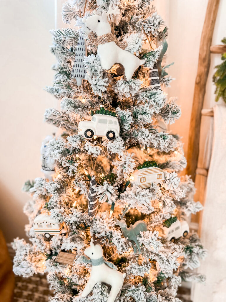 In a child bedroom, there is a small flocked Christmas tree in a woven basket, with a rattan star on top. There are several different types of ornaments: an old white station wagon, a white stuffed animal deer, and a blue felt deer.