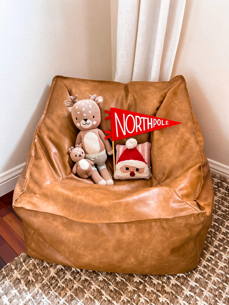 In a child bedroom, there is a Pottery Barn Teen leather ottoman sitting pouf on the floor. On the ottoman is a Cuddle and Kind deer and a small baby Cuddle and Kind doll deer. There is a medium sized red pennant flag with white lettering that says "North Pole." There is also a mini square pillow with a Santa face knitted on the front.