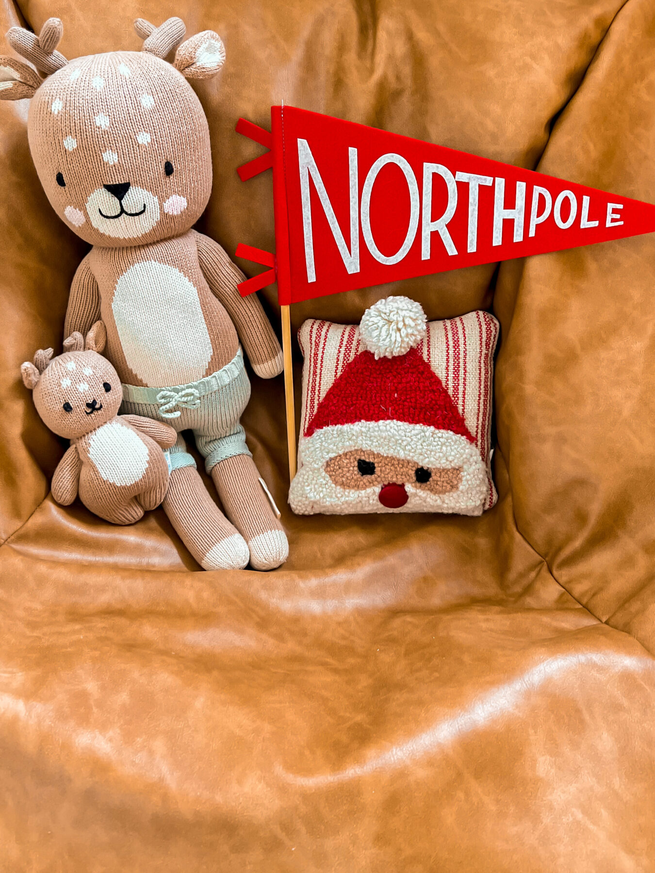 In a child bedroom, there is a Pottery Barn Teen leather ottoman sitting pouf on the floor. On the ottoman is a Cuddle and Kind deer and a small baby Cuddle and Kind doll deer. There is a medium sized red pennant flag with white lettering that says "North Pole." There is also a mini square pillow with a Santa face knitted on the front.