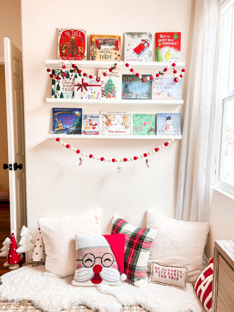 This is a child bedroom decorated for Christmas. There is sitting area on the floor with several Christmas pillows and above that is three bookshelves. The bookshelves have Christmas and Holiday children's books, and is decorated with red and white pom pom Christmas inspired garland.